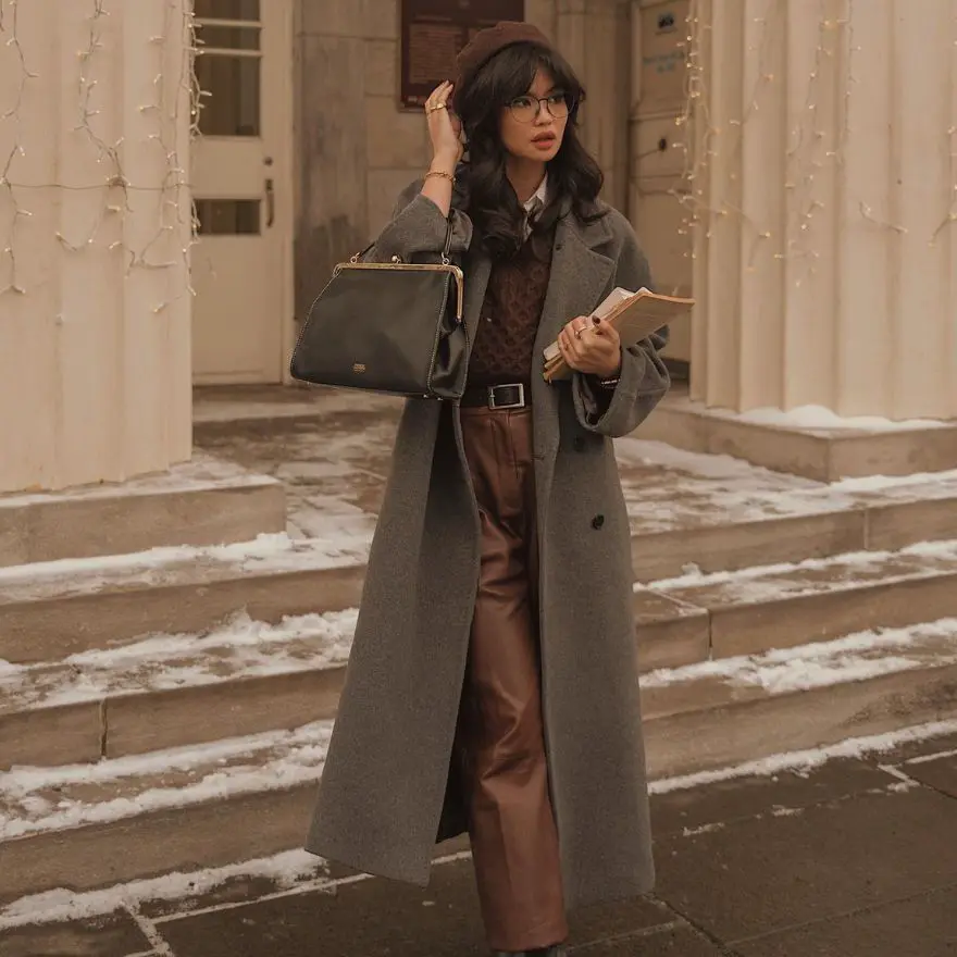 Person in dark academia winter outfit with gray coat, brown beret, and leather pants holding stack of books on snowy steps
