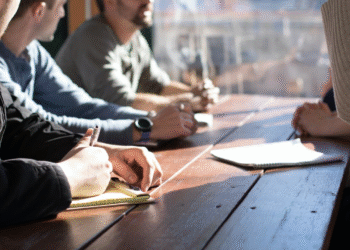 Hands and notebooks around a wooden table in a collaborative meeting, with people writing and discussing ideas.