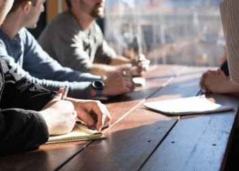 Hands and notebooks around a wooden table in a collaborative meeting, with people writing and discussing ideas.