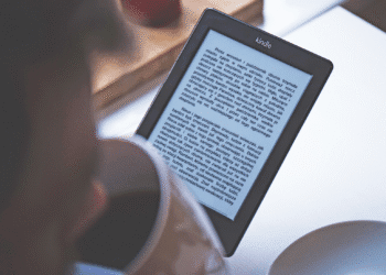 Person reading an ebook on a Kindle e-reader device while holding a cup of coffee in a cozy setting